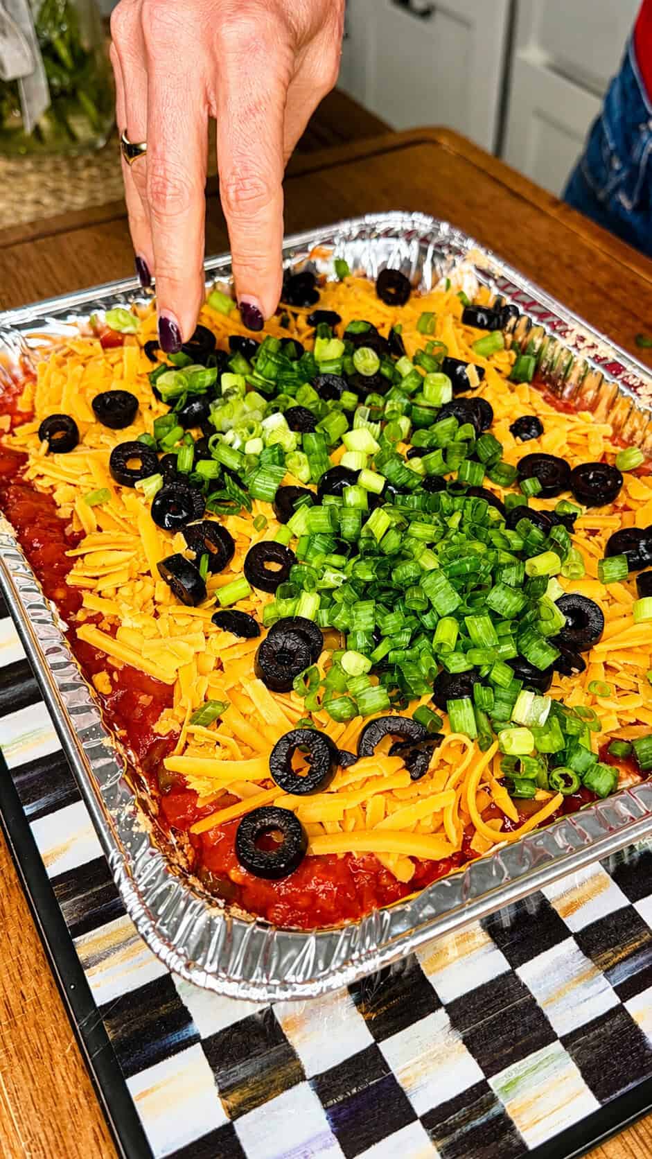 A hand sprinkles chopped green onions onto a layered dip topped with shredded cheddar cheese and sliced black olives, in a rectangular foil tray on a checkered surface.