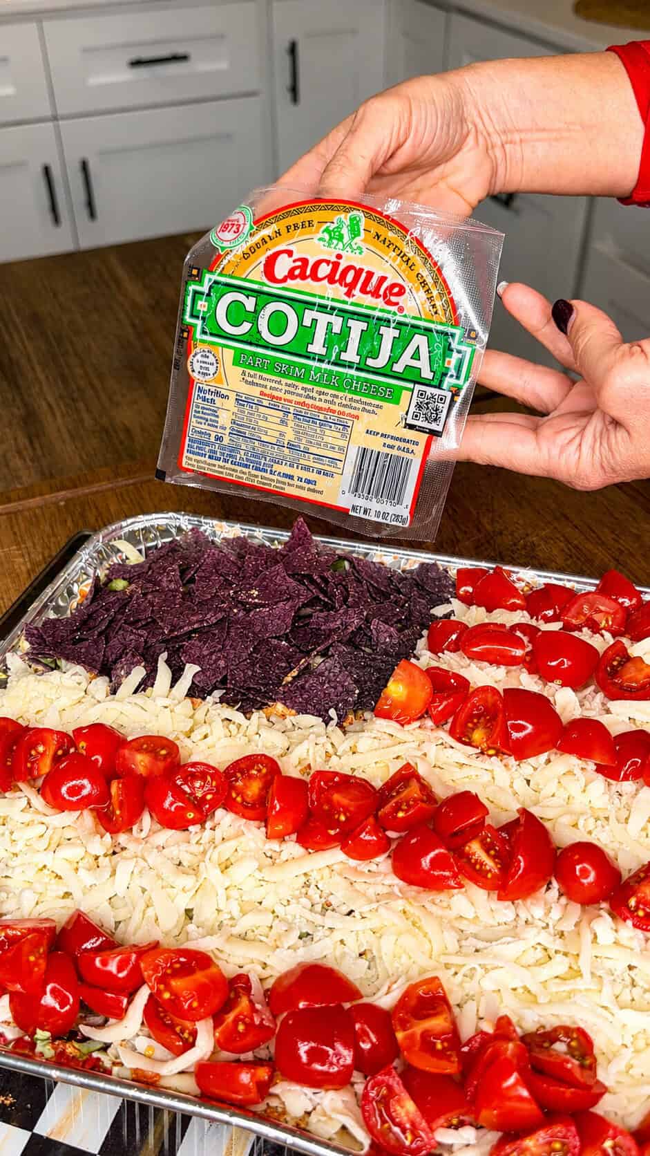 A hand holds a package of Cacique Cotija cheese above a tray of shredded cheese, cherry tomatoes, and blue corn chips arranged in a pattern on a baking sheet in a kitchen.