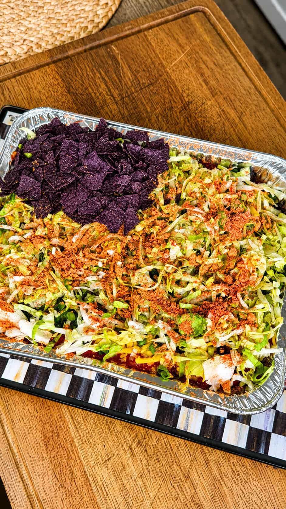 A tray of layered taco salad topped with shredded lettuce, crushed tortilla chips, and seasoning, with a pile of blue corn tortilla chips on one side, set on a wooden surface.