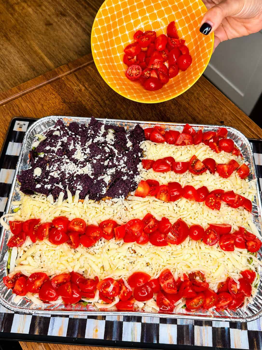 A person sprinkles chopped cherry tomatoes onto a tray of dip arranged to resemble the American flag, with tomato stripes, shredded cheese, and blue tortilla chips topped with cheese for the stars.
