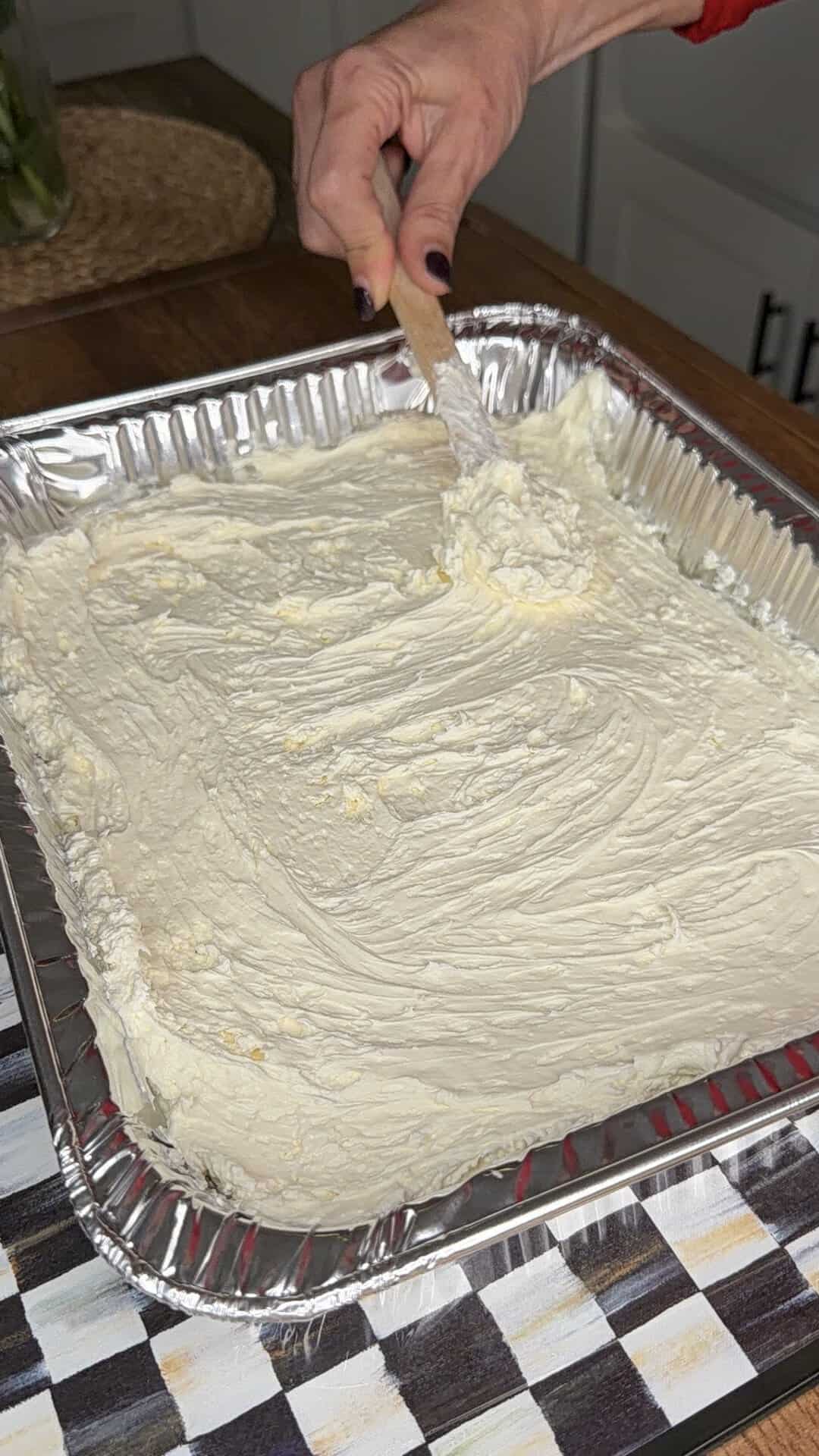 A hand holding a spatula spreads creamy batter evenly in a large rectangular aluminum baking pan on a kitchen countertop.