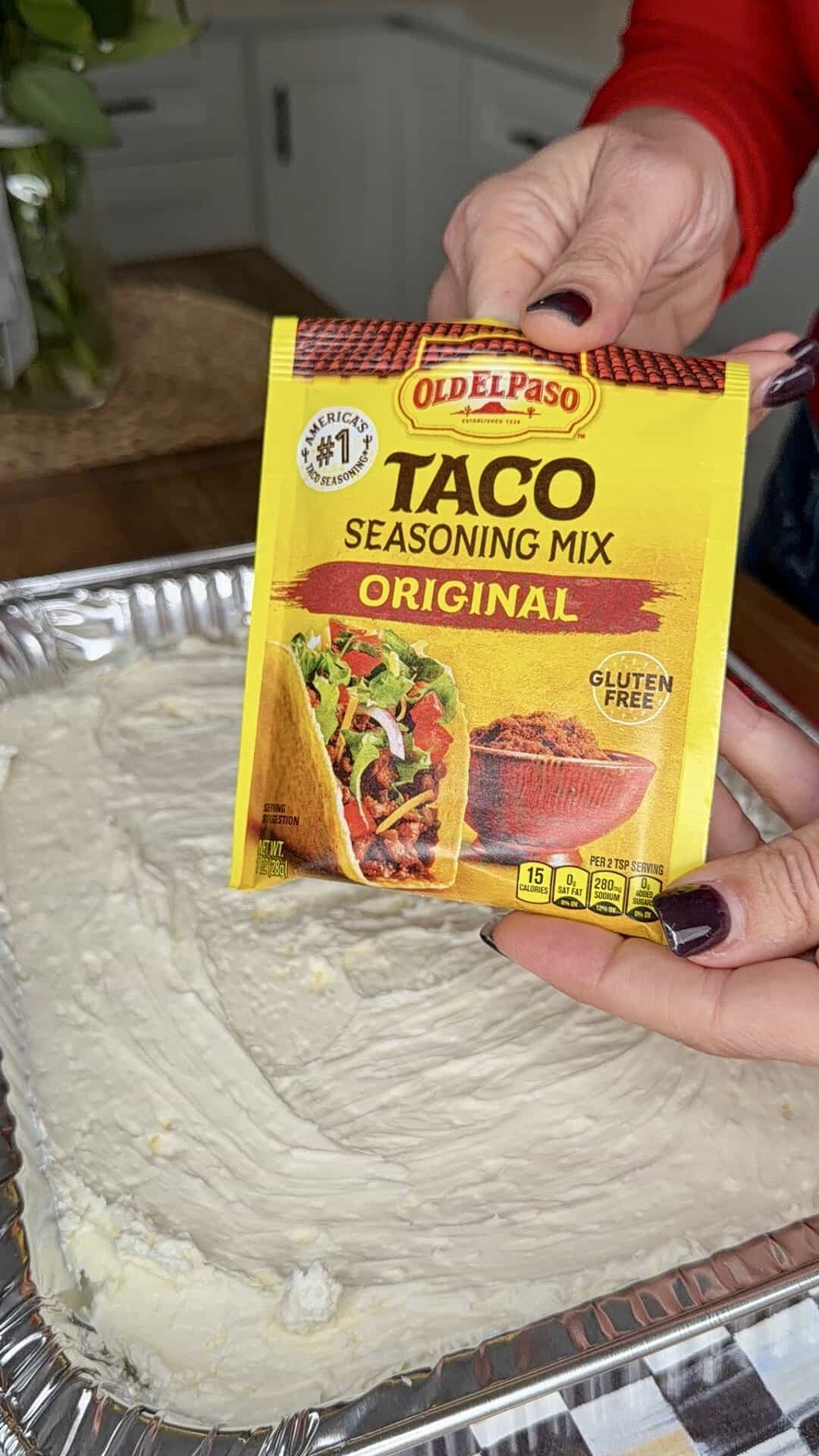 A person holds a packet of Old El Paso Taco Seasoning Mix over a foil tray filled with a creamy white mixture in a kitchen setting.
