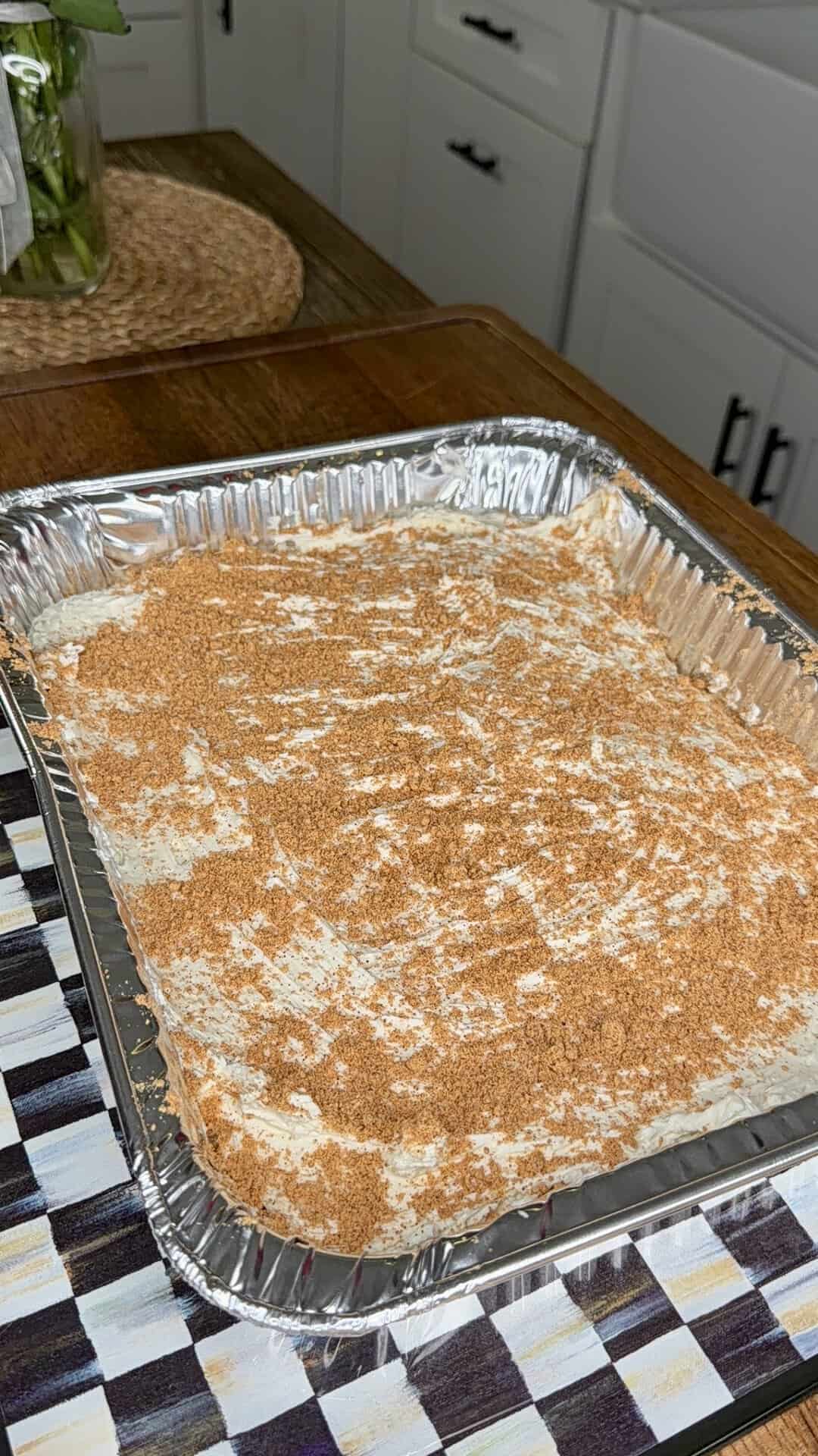 A large rectangular aluminum tray contains a dessert topped with a light layer of cinnamon or brown sugar. The tray sits on a patterned placemat on a wooden table in a kitchen setting.