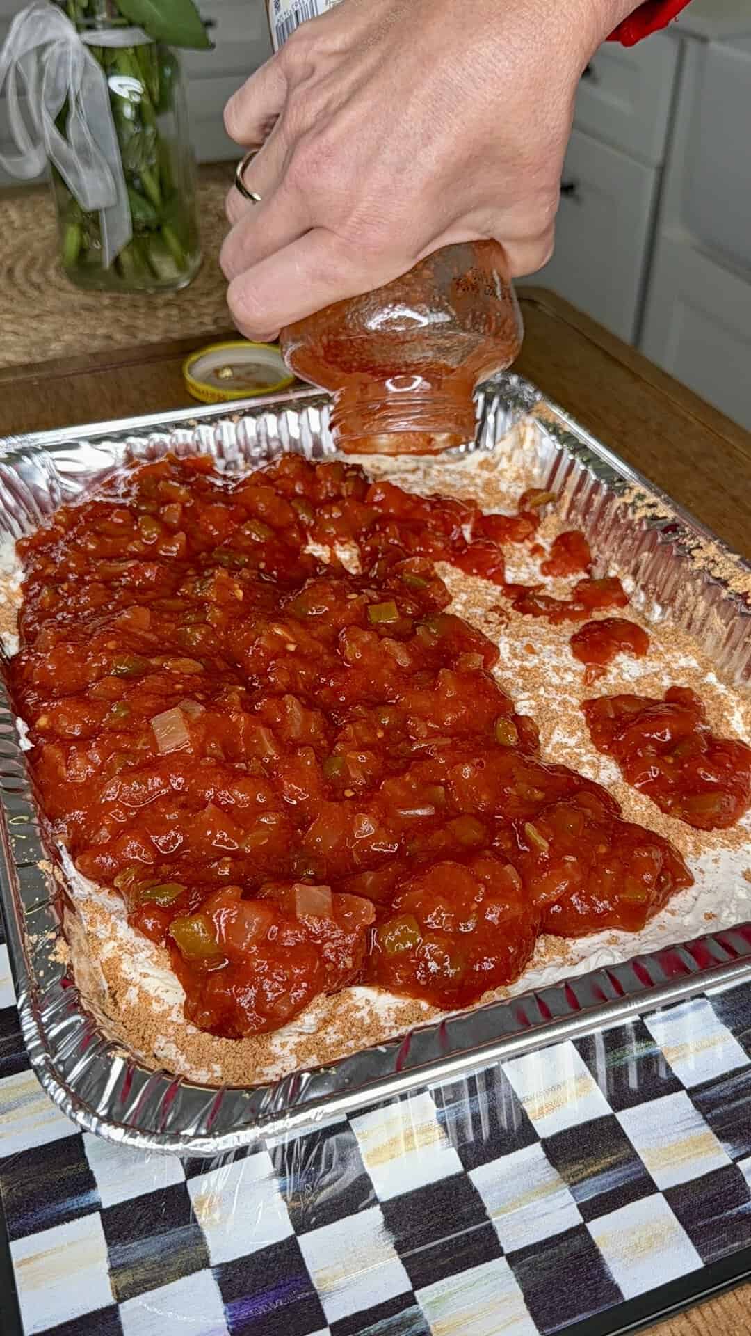 A hand pours chunky tomato sauce from a glass jar over a layered mixture in a disposable aluminum baking pan on a kitchen counter.