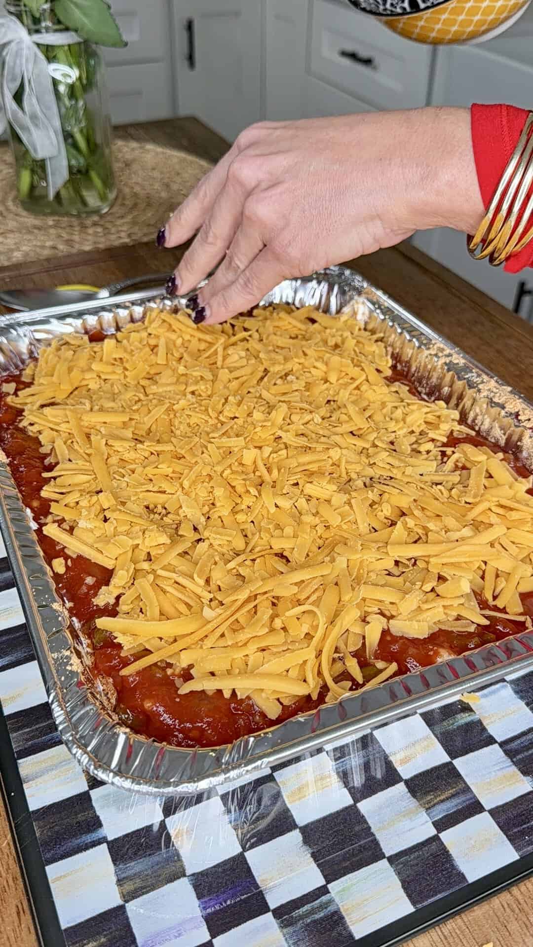 A hand sprinkles shredded cheddar cheese over a casserole in a foil tray on a kitchen counter. The casserole is topped with tomato sauce and is being prepared for baking.