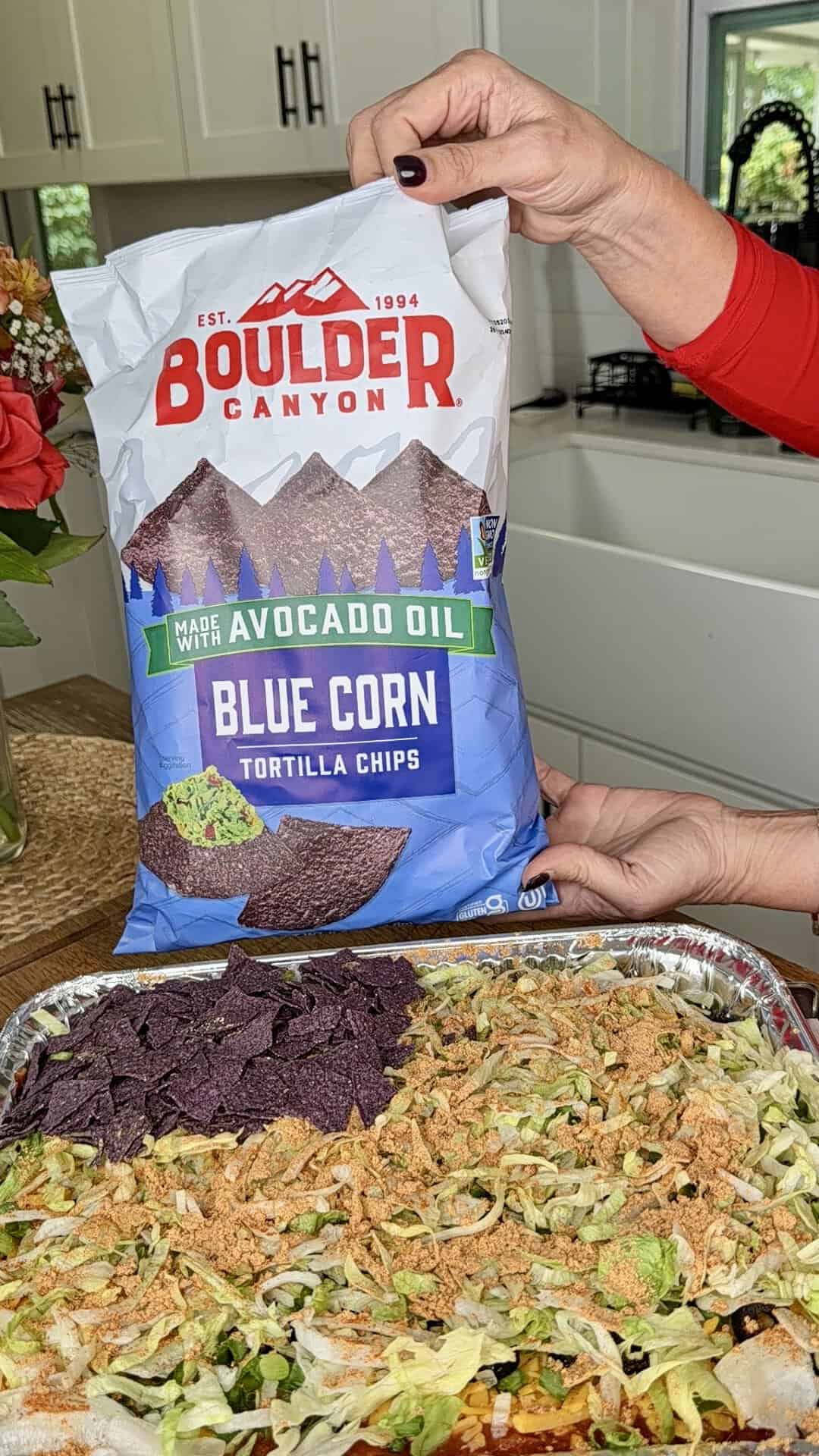A person holds a bag of Boulder Canyon Avocado Oil Blue Corn Tortilla Chips above a tray of salad topped with shredded lettuce and crispy toppings in a kitchen.