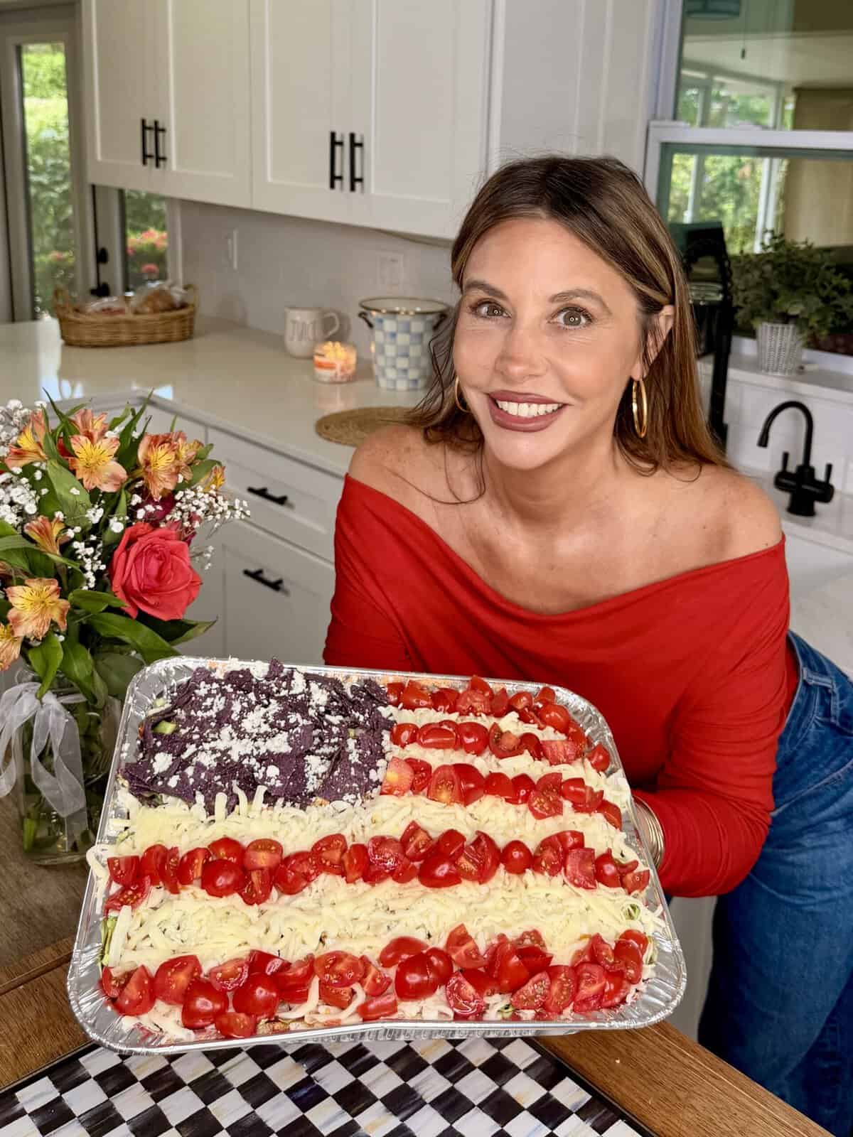 A woman in a red off-shoulder top smiles while holding a tray of food decorated like the American flag, with cheese, tomatoes, and a dark topping. She stands in a bright kitchen next to a vase of flowers.