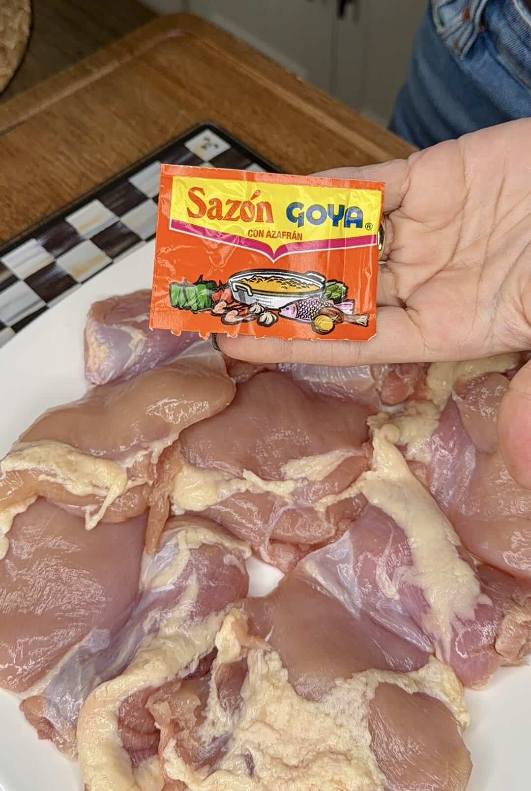 A hand holds a packet of Sazón Goya seasoning over a plate with several pieces of raw chicken, ready to be seasoned.
