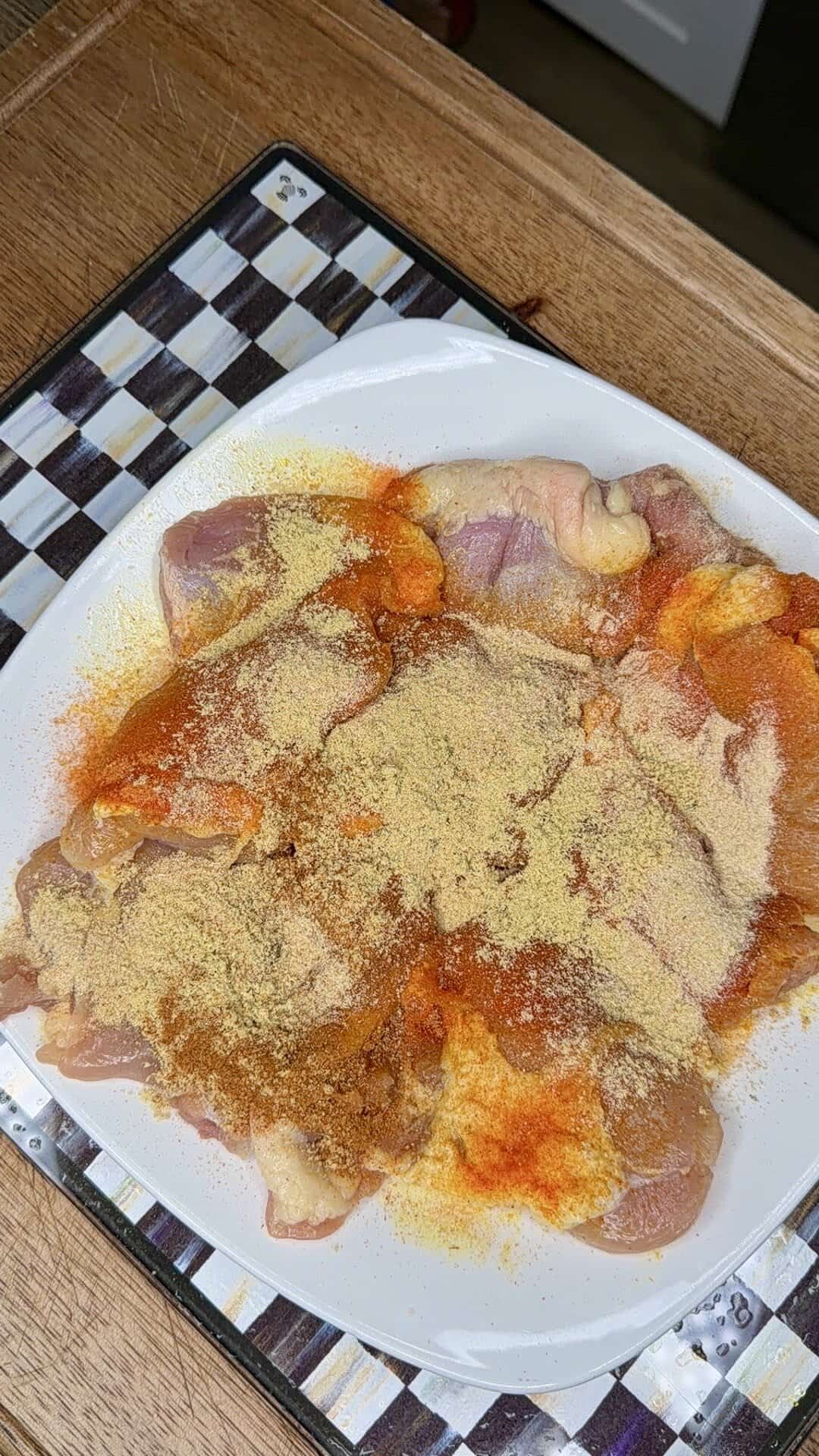 Raw chicken pieces on a white plate, sprinkled with various spices, including paprika and a yellowish powder, placed on a checkered placemat on a wooden table.