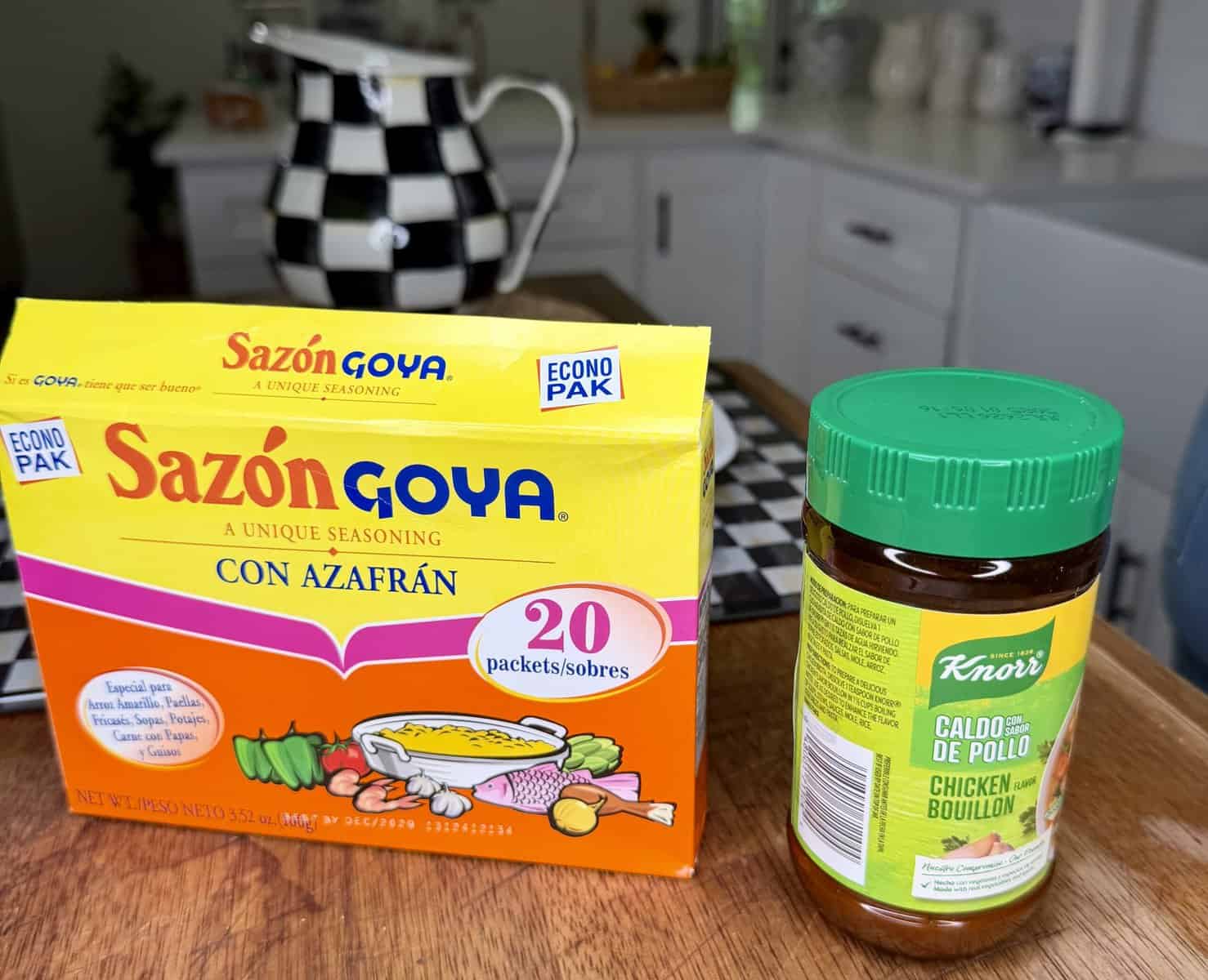 A box of Sazón Goya seasoning and a jar of Knorr chicken bouillon sit on a wooden kitchen counter, with a black-and-white checkered teapot and matching items in the background.