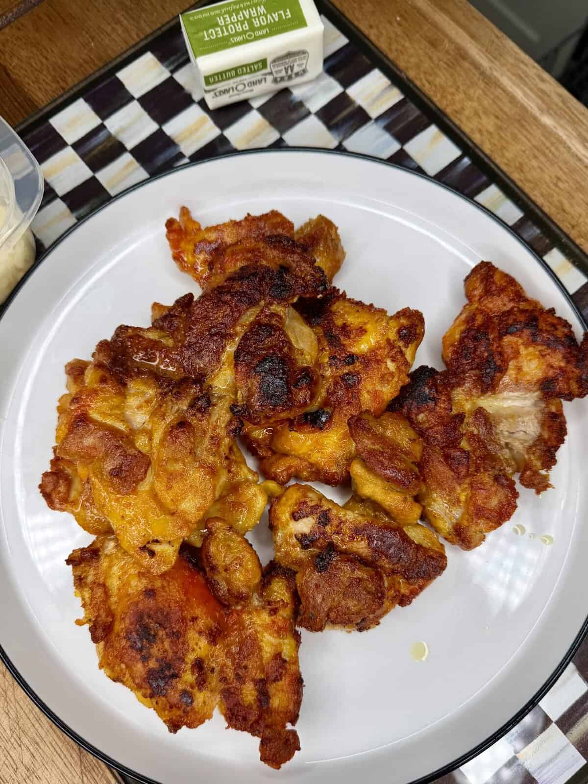 A white plate with several pieces of browned, cooked chicken thighs on a checkered placemat. A small packet of salted butter is visible in the background.