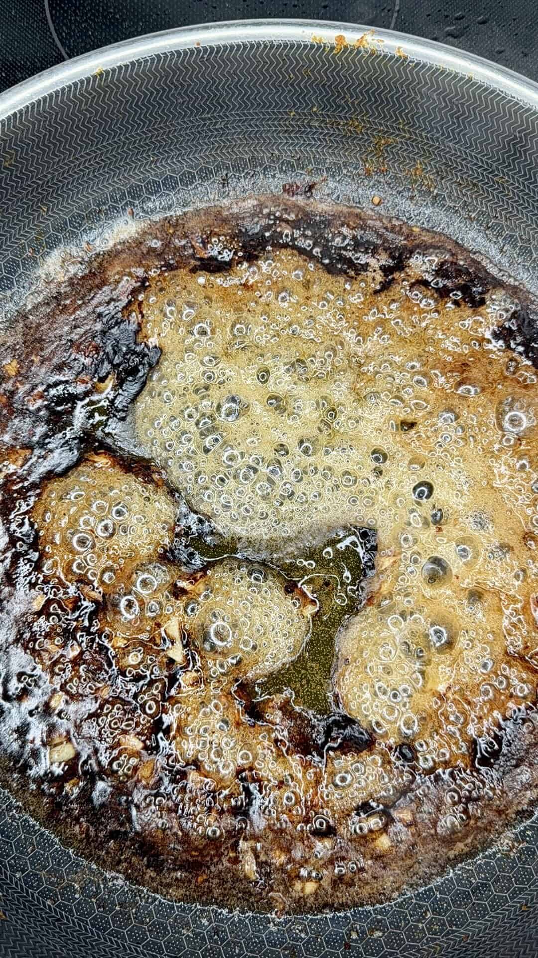 Caramelized sugar bubbling and turning brown in a frying pan, with some areas darker and slightly burnt, creating a mix of golden and deep brown textures.