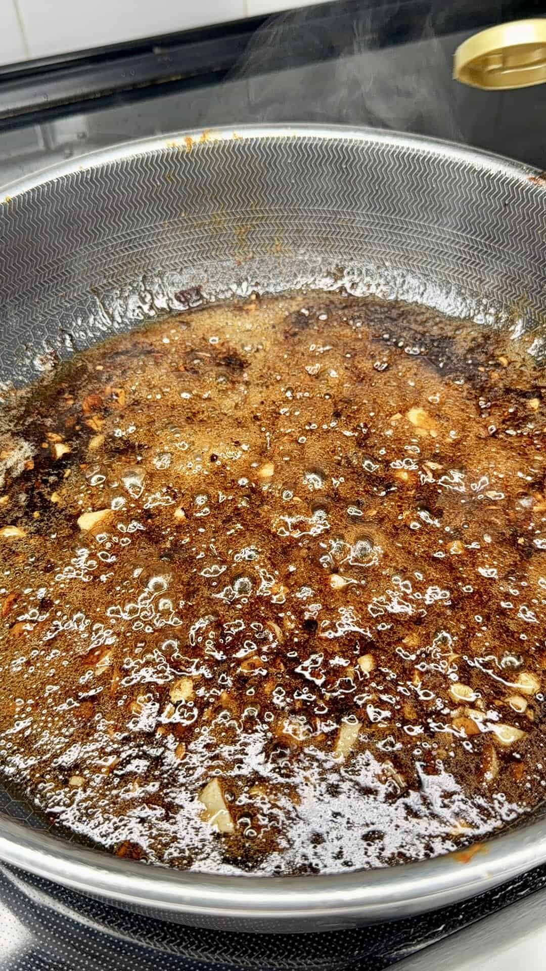 A close-up of a frying pan on a stovetop containing bubbling oil with chopped garlic, chili flakes, and spices, creating a rich, aromatic sauce. Steam is rising from the surface.