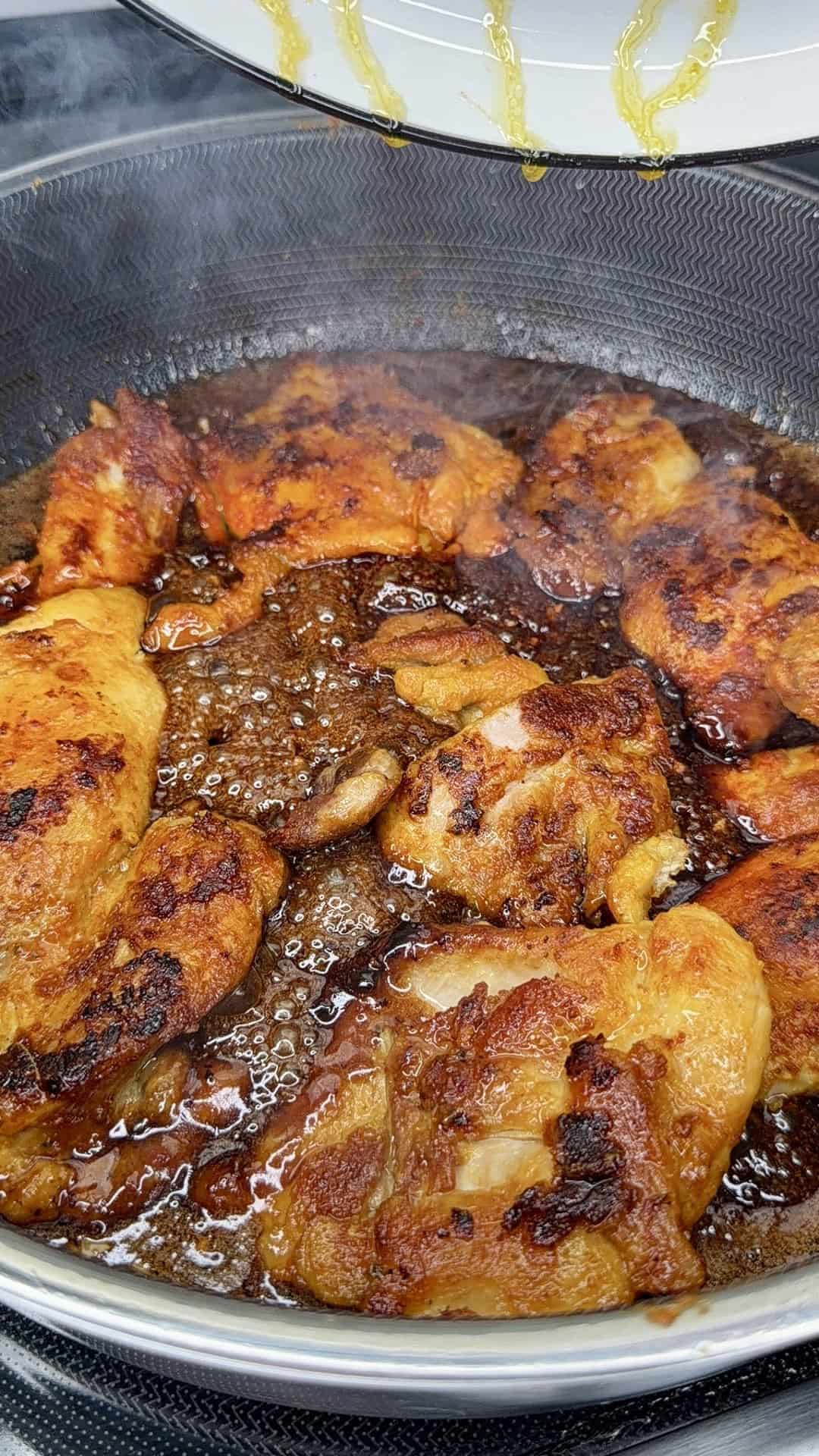Golden-brown chicken pieces sizzling in a frying pan with bubbling oil, some steam rising from the hot surface, and sauce residue visible on the pan and the nearby lid.