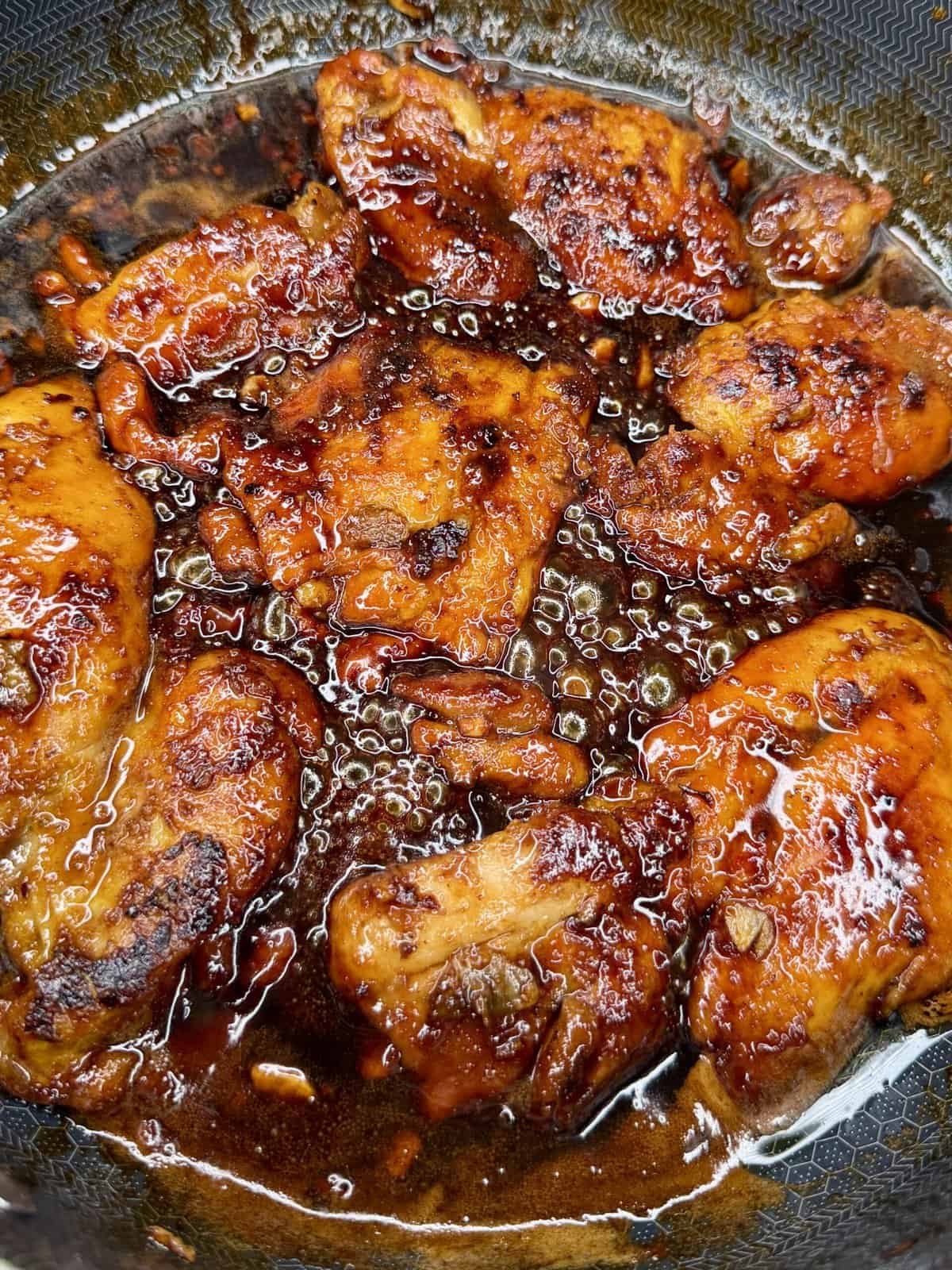 Chicken pieces are simmering in a pan, coated in a rich, glossy, dark brown sauce with visible garlic and seasoning, giving the dish a caramelized and flavorful appearance.
