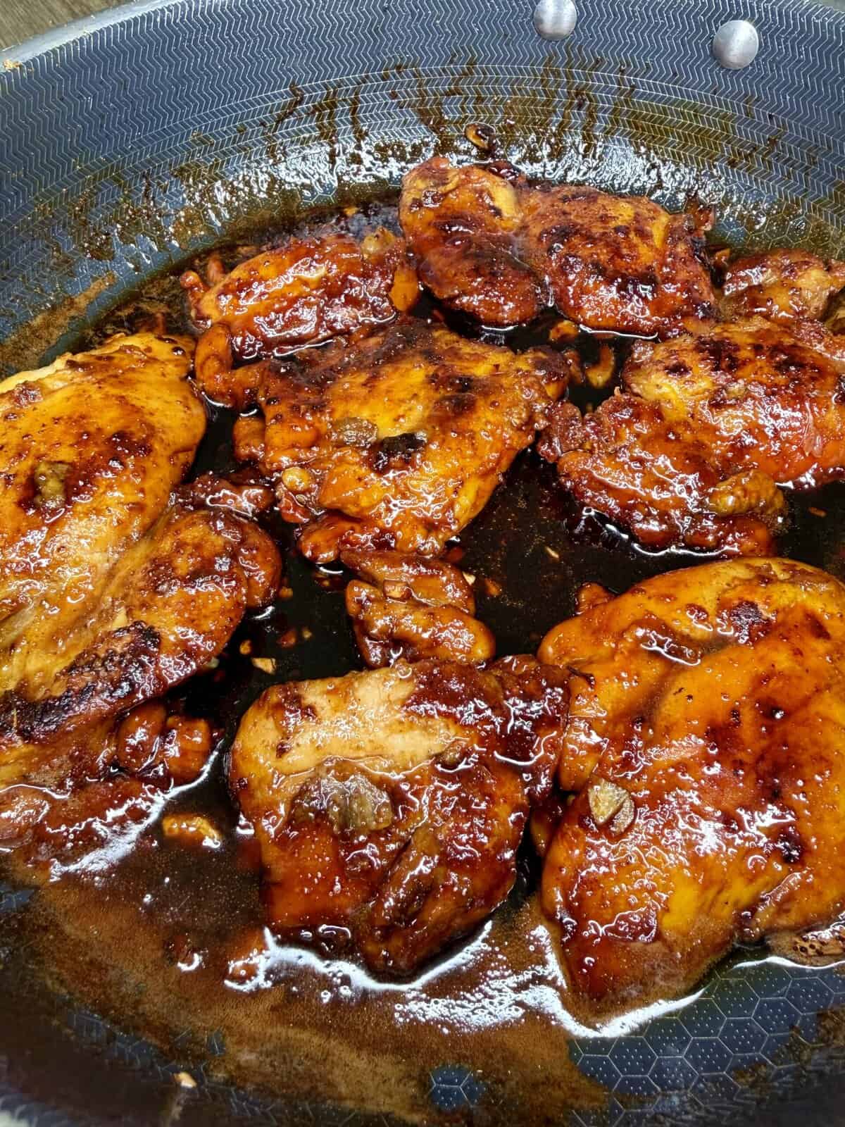 Chicken thighs coated in a glossy, caramelized sauce cook in a skillet. The sauce is thick, dark, and sticky, clinging to the browned chicken pieces, with oil pooling around them in the pan.