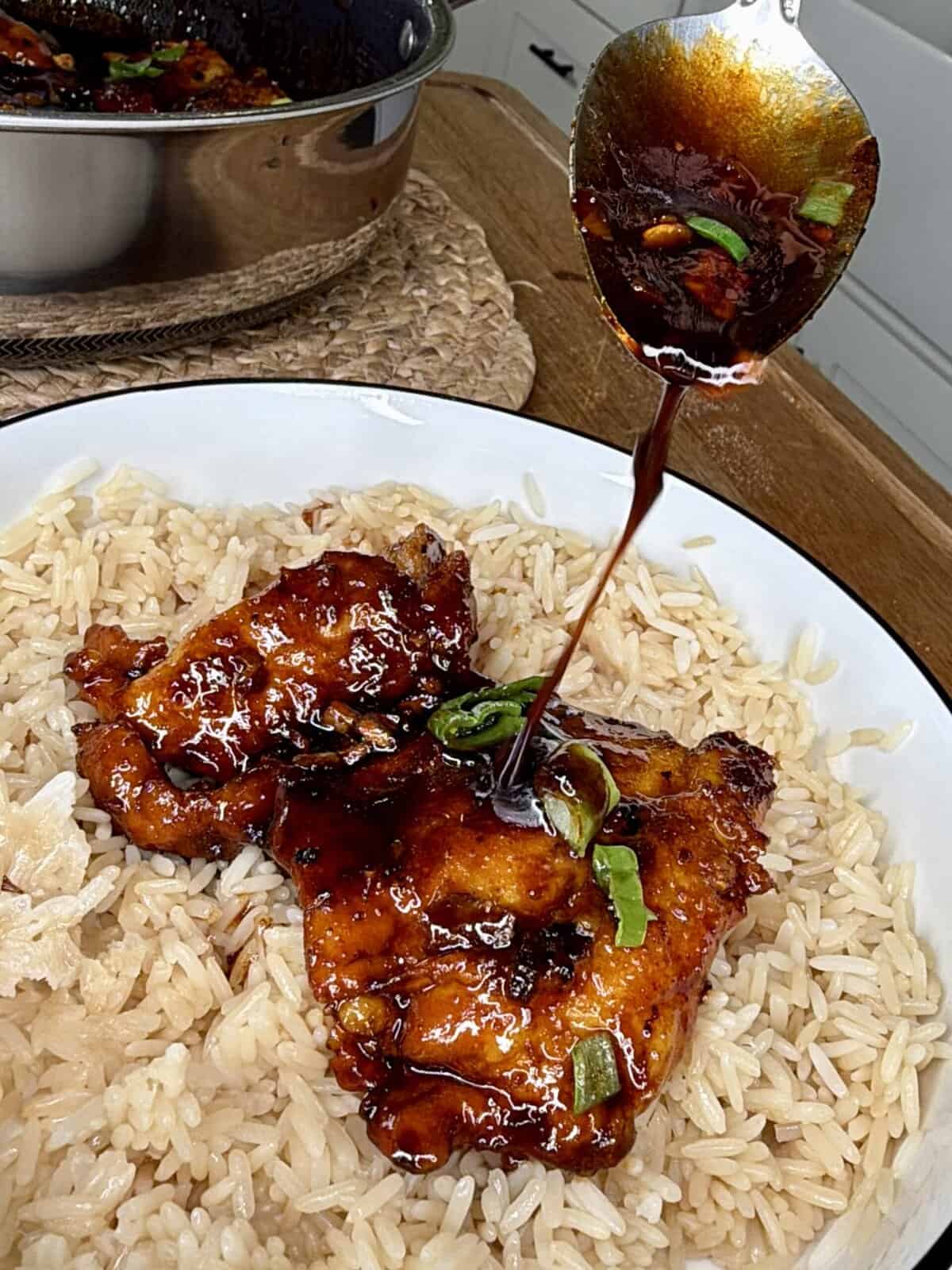 A plate of rice topped with two glazed chicken thighs is shown, while a spoon drizzles dark sauce with green onions over the chicken. A pot with more chicken is visible in the background.