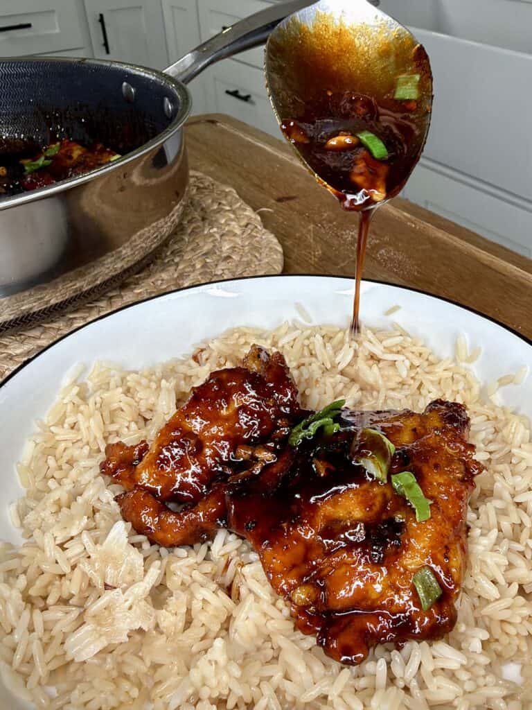 A plate of cooked white rice topped with glazed chicken and green onions, while a spoon drizzles dark sauce over the chicken. A pan with more sauce is visible in the background on a wooden surface.