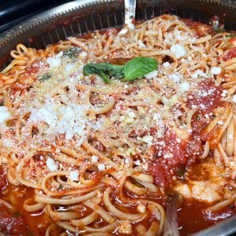 A close-up of spaghetti topped with tomato sauce, grated Parmesan cheese, crumbled cheese, and a fresh basil garnish served in a large metal dish.