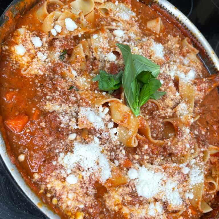 A close-up of wide pasta noodles in a rich tomato meat sauce, topped with grated cheese and a sprig of fresh basil in the center.