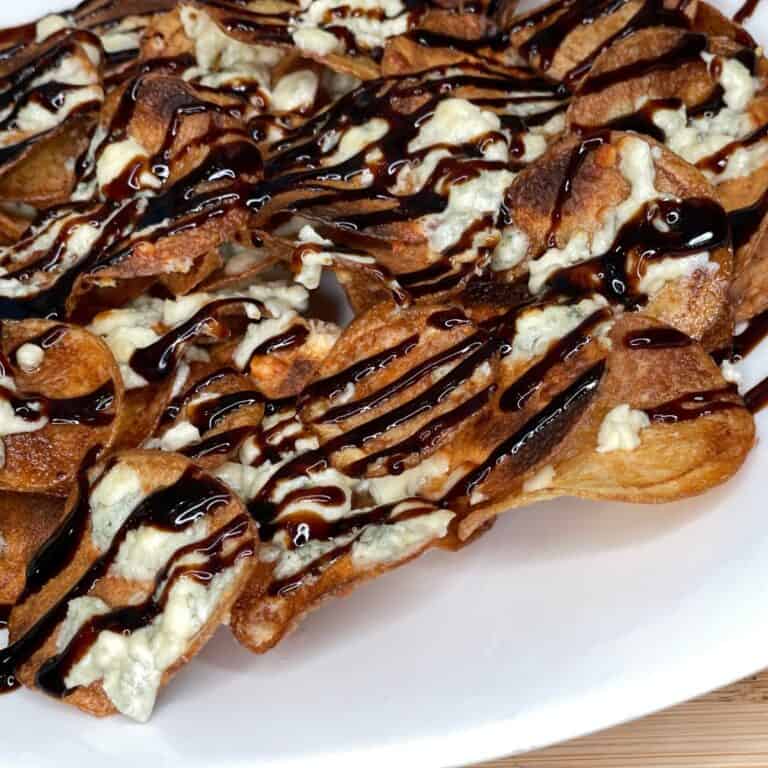 A plate of crispy potato chips topped with melted blue cheese and drizzled with a dark balsamic glaze, served on a white dish.