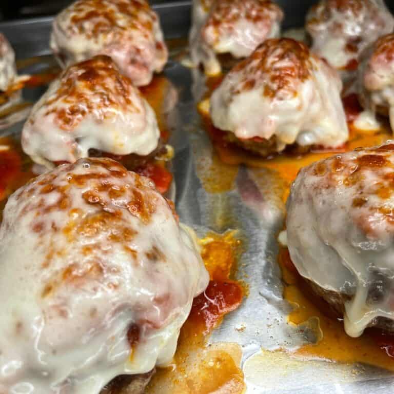 Mini meatloaves topped with melted, slightly browned mozzarella cheese and tomato sauce on a baking sheet.