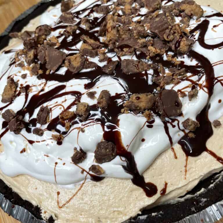A close-up of a creamy pie topped with whipped cream, chocolate syrup drizzle, and chunks of chocolate candy, in a dark cookie crust.