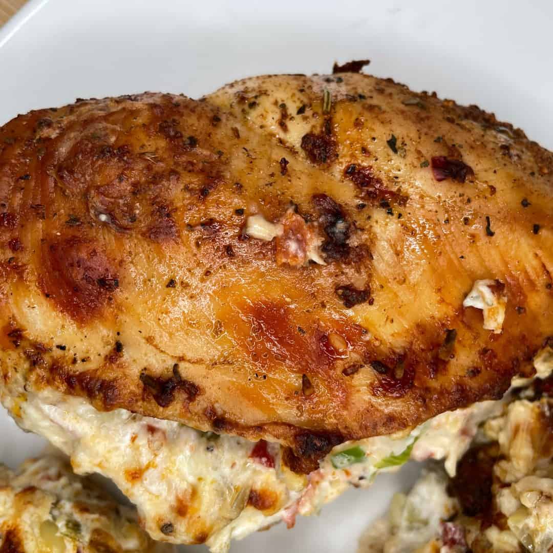 A close-up of a baked chicken breast stuffed with a creamy mixture, seasoned with herbs and spices, served on a white plate. Some of the filling is visible where the chicken is slightly open.