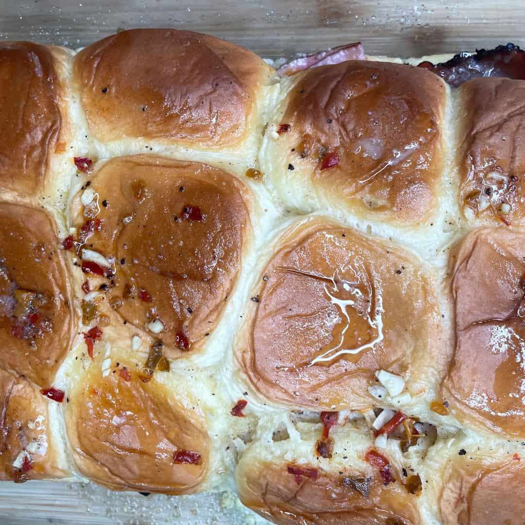 A close-up of golden, baked slider buns brushed with melted butter, minced garlic, and red pepper flakes, with some sandwich filling visible at the edges on a wooden surface.