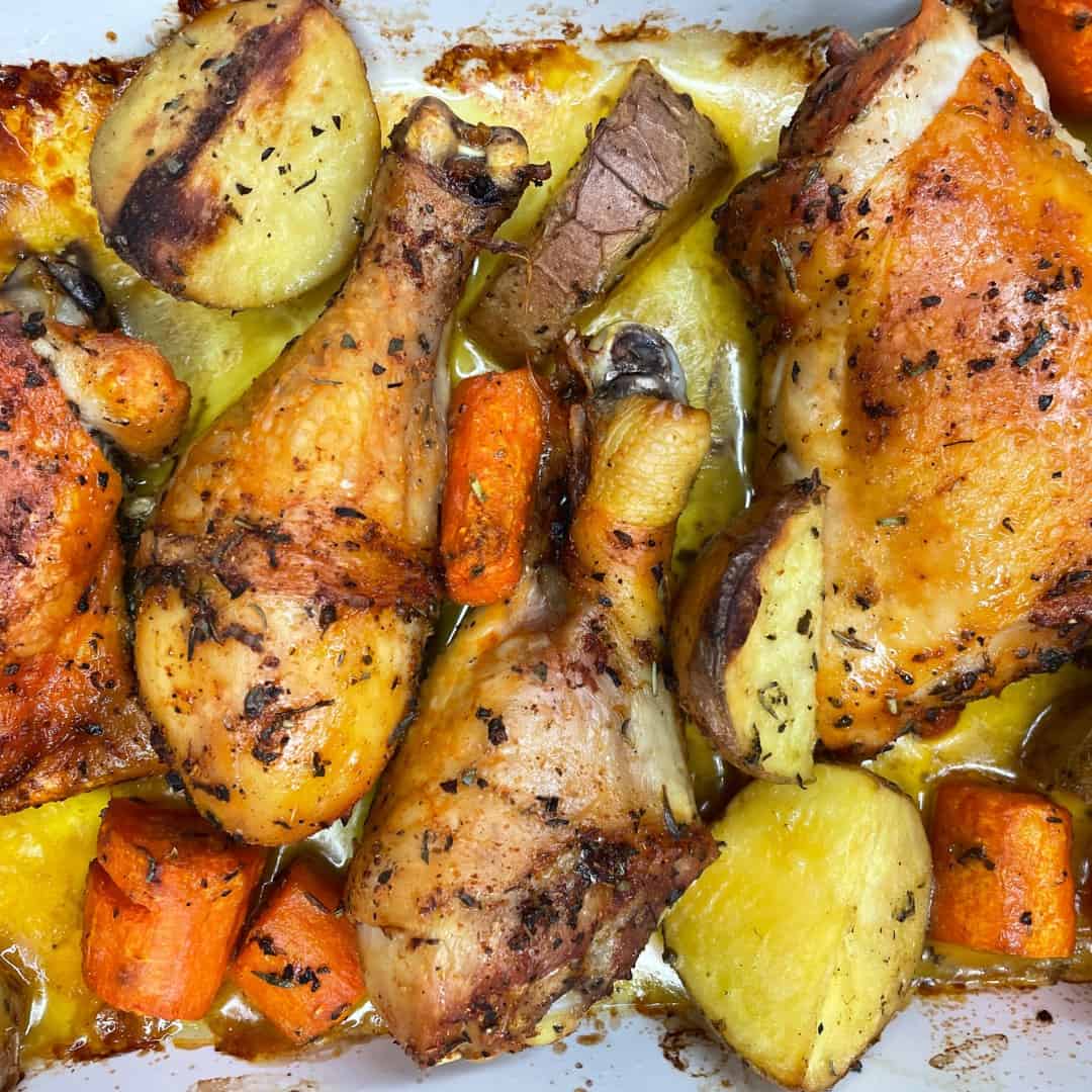 Roasted chicken drumsticks with golden brown skin, surrounded by chunky pieces of potatoes and carrots, all seasoned with herbs and cooked in a baking dish with visible juices.