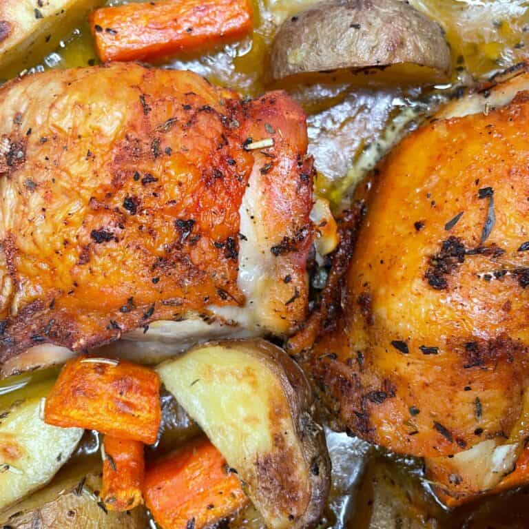 Close-up of roasted chicken thighs with crispy skin, surrounded by golden-brown potato wedges and chunks of carrot, all seasoned and cooked together.