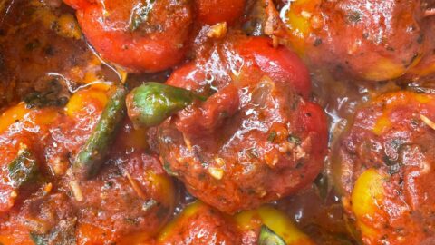 Six stuffed bell peppers covered in a rich tomato sauce, garnished with herbs, simmering in a pot. The peppers are red and yellow, with visible chunks of filling inside.