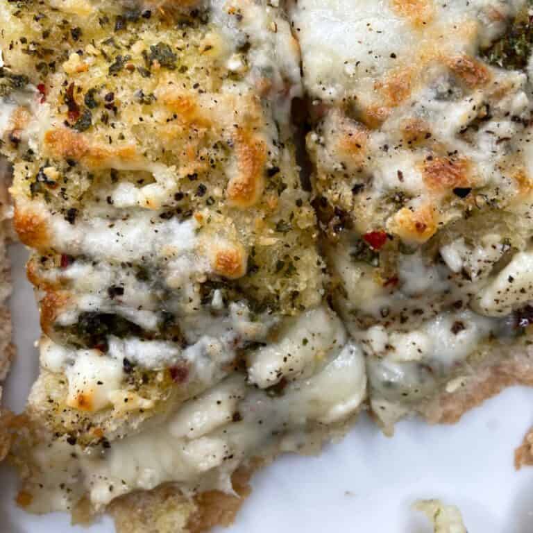 Close-up of two slices of cheesy garlic bread topped with melted cheese, herbs, and cracked black pepper, showing a golden-brown, bubbly texture on a white plate.