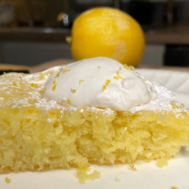 A close-up of a moist lemon cake topped with a dollop of whipped cream and lemon zest, with a whole lemon in the background.