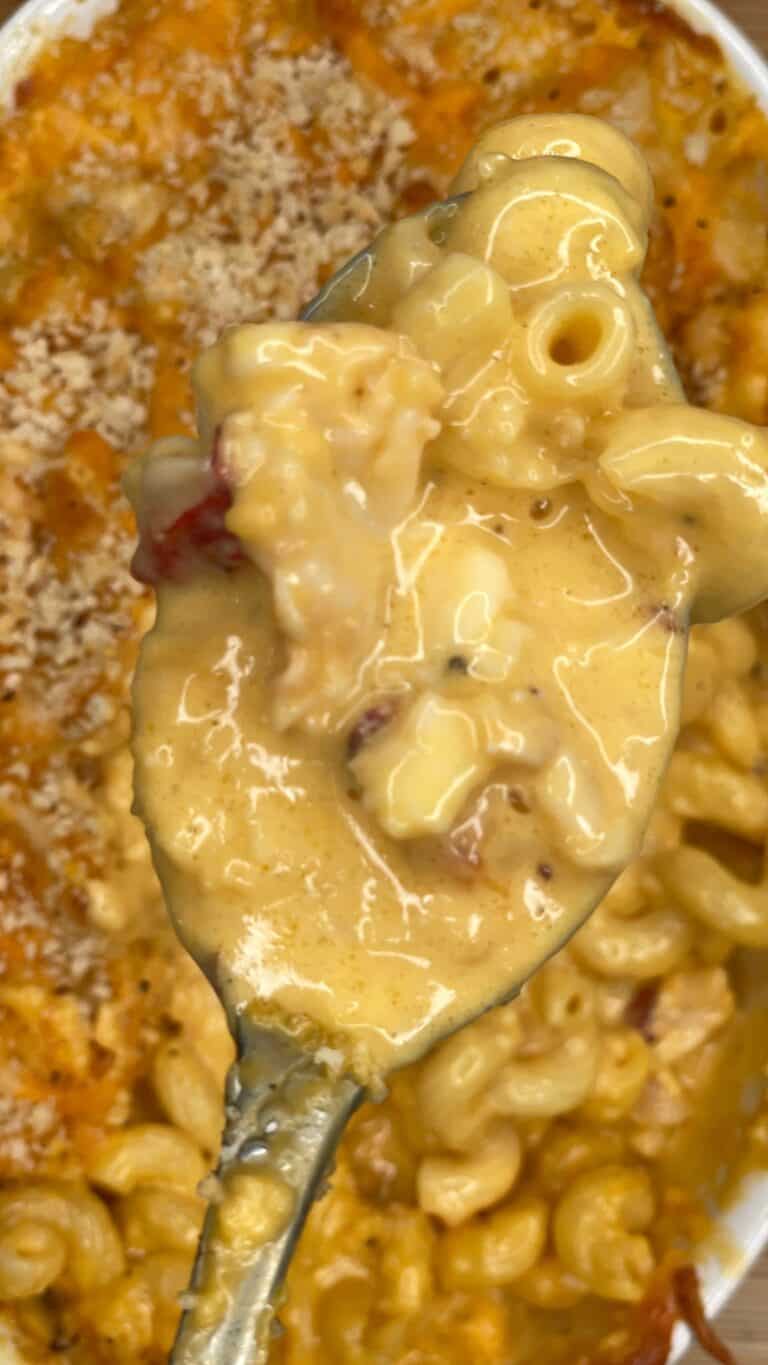 A close-up of a spoonful of creamy macaroni and cheese with bits of bacon, held above a baking dish filled with more cheesy macaroni.