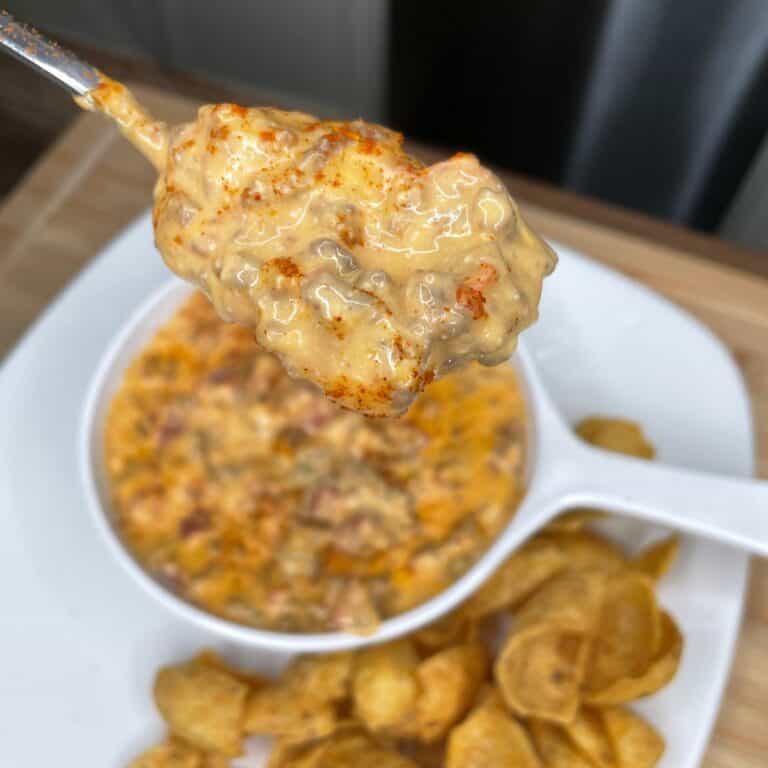 A close-up of a spoonful of creamy, cheesy dip with visible bits of meat, held above a bowl of the same dip. Potato chips are on the side of the white plate.