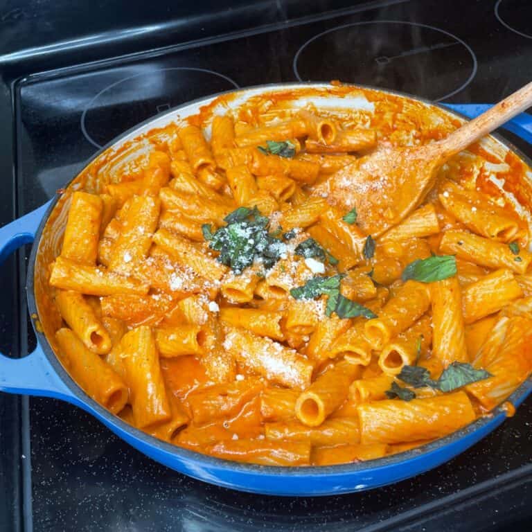A blue pan filled with creamy rigatoni pasta in tomato sauce, topped with fresh basil leaves and grated cheese. A wooden spoon rests in the pasta. The pan sits on a black stovetop.