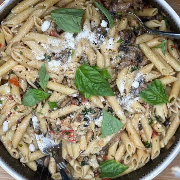 A close-up of a pan filled with penne pasta mixed with tomatoes, mushrooms, herbs, grated cheese, and garnished with fresh basil leaves. A fork and spoon are resting in the pasta.