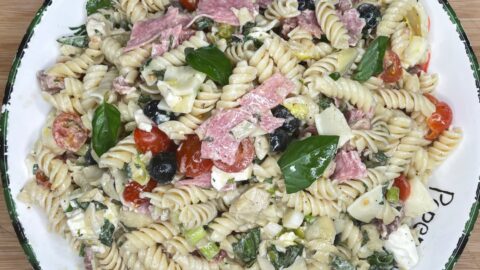 A bowl of rotini pasta salad with cherry tomatoes, black olives, salami slices, artichoke hearts, feta cheese, fresh basil, and a creamy dressing, served on a white plate with a decorative rim.