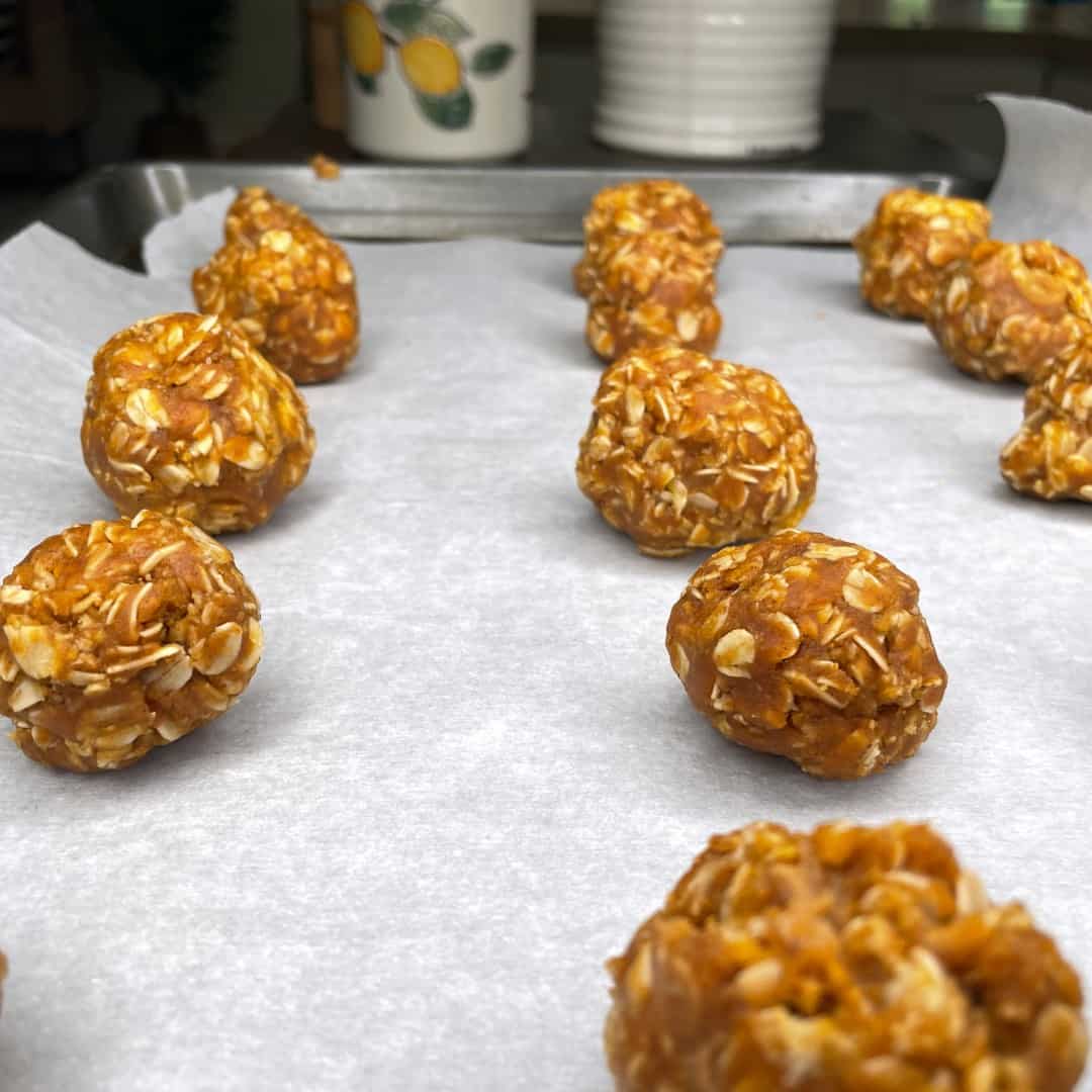 Several round, orange oat balls are arranged on a parchment-lined baking sheet, ready to be baked. The background features a lemon-patterned container and stacked white dishes.