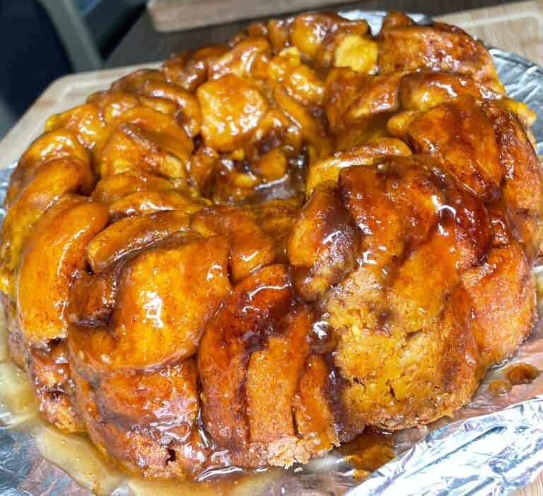 A close-up of a freshly baked monkey bread, golden brown and sticky with cinnamon sugar glaze, sitting on a foil-covered surface. The bread appears soft and gooey with visible sweet syrup.