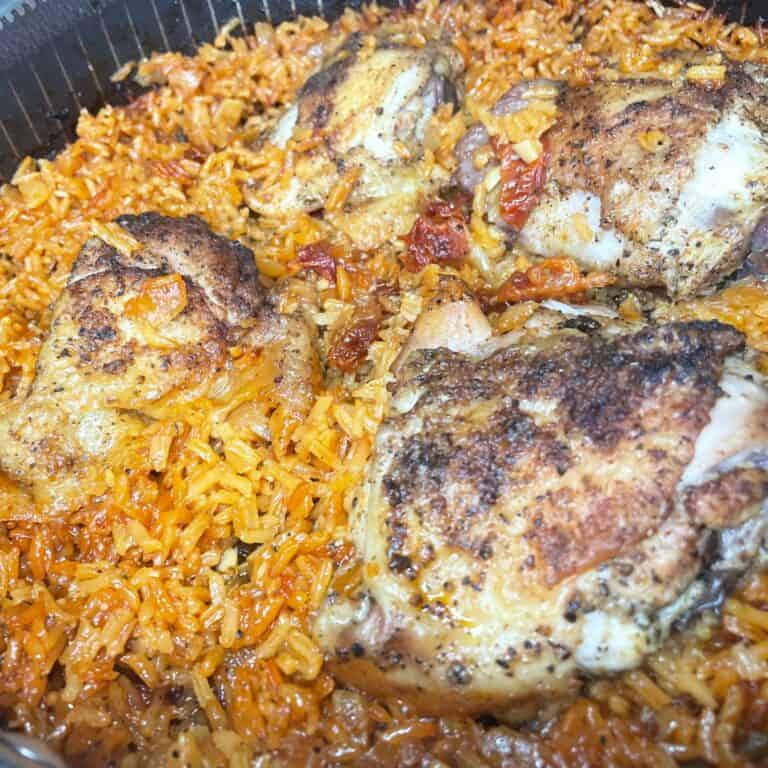 Four seasoned, browned chicken thighs sit atop a bed of orange-yellow rice mixed with small bits of tomato, all cooked together in a large, deep pan.
