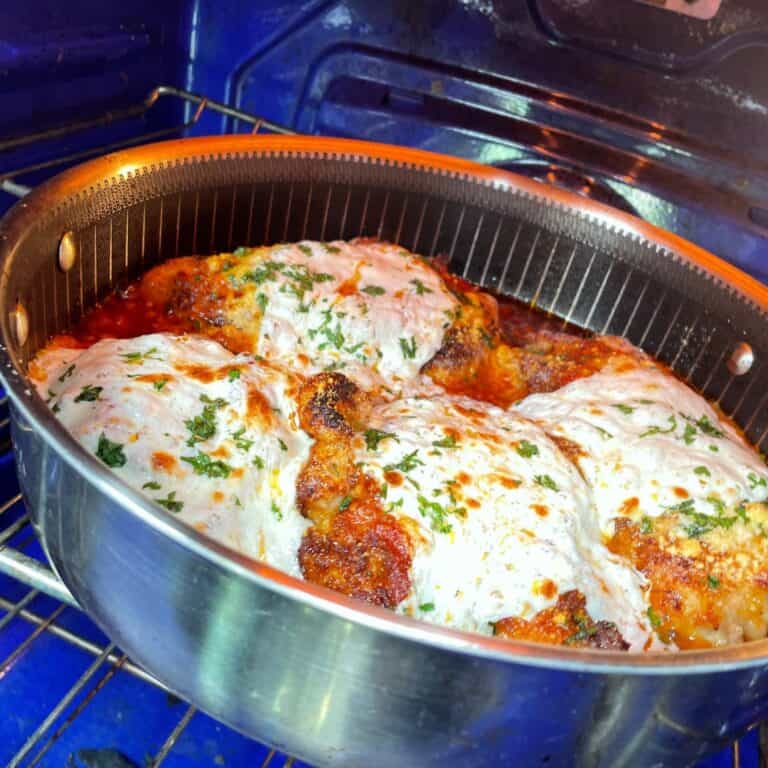 A round metal pan in an oven holds breaded chicken breasts topped with melted mozzarella cheese and sprinkled with chopped parsley, likely chicken parmesan, baking to a golden brown.