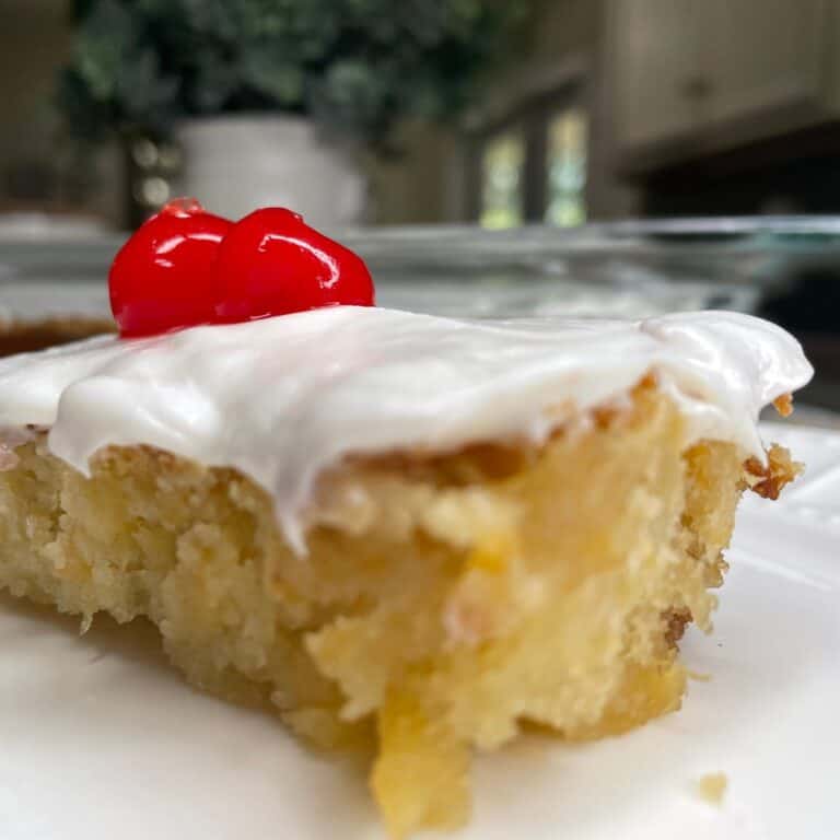 A close-up of a slice of frosted yellow cake topped with two bright red cherries, placed on a white plate with a blurred kitchen background.