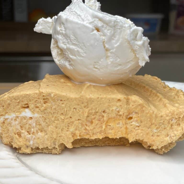 A slice of creamy pumpkin cheesecake with a graham cracker crust, topped with a large dollop of whipped cream, sits on a white plate. A bite has been taken from the cheesecake.