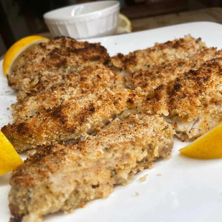 Golden-brown, breaded fish fillets arranged on a white plate, garnished with lemon wedges. A small white dipping bowl is blurred in the background.