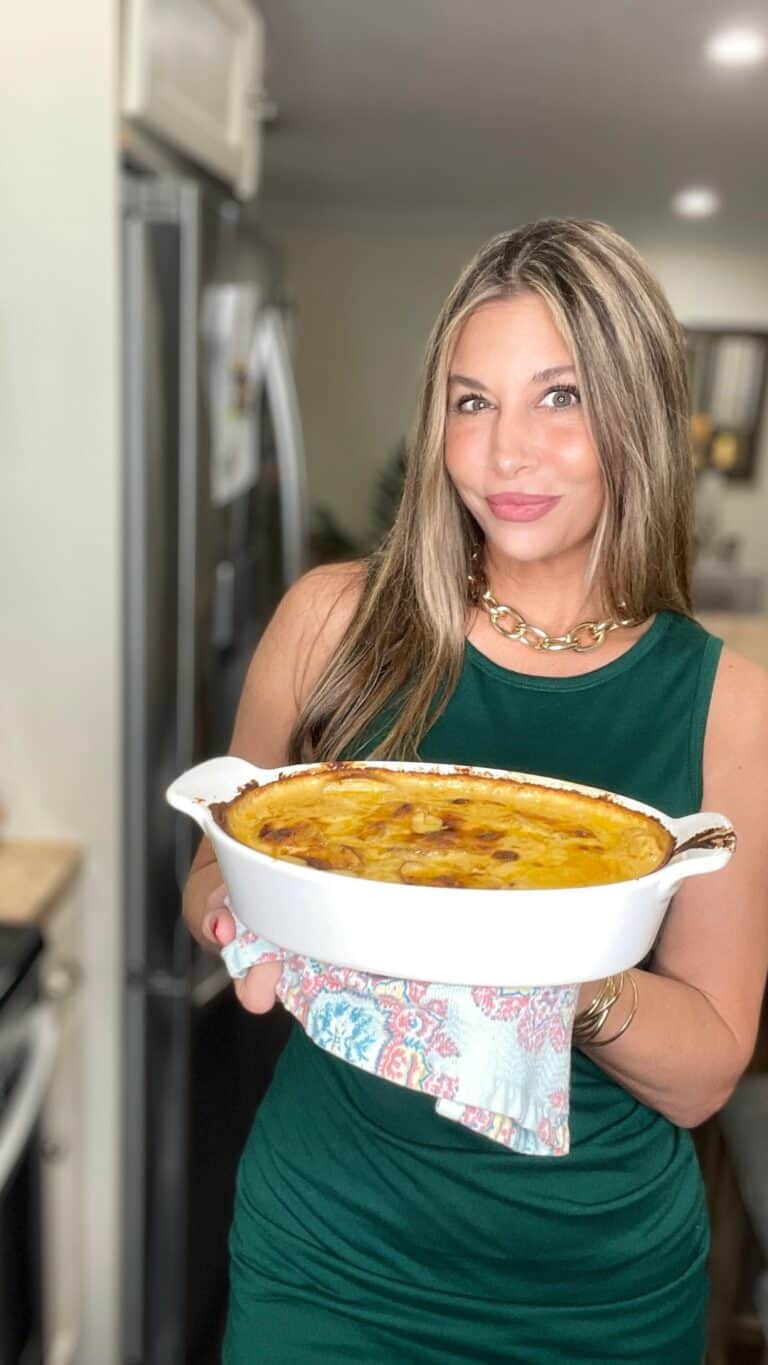 A woman in a green dress holds a baked casserole in a white dish, smiling in a kitchen setting.