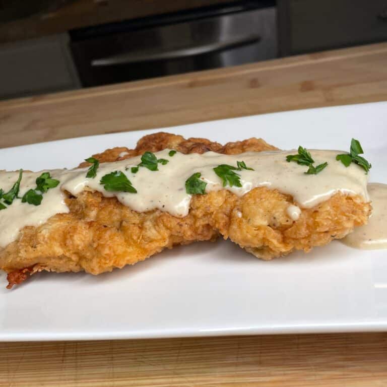 Two pieces of crispy fried chicken topped with creamy white gravy and garnished with chopped parsley, served on a white rectangular plate on a wooden surface.