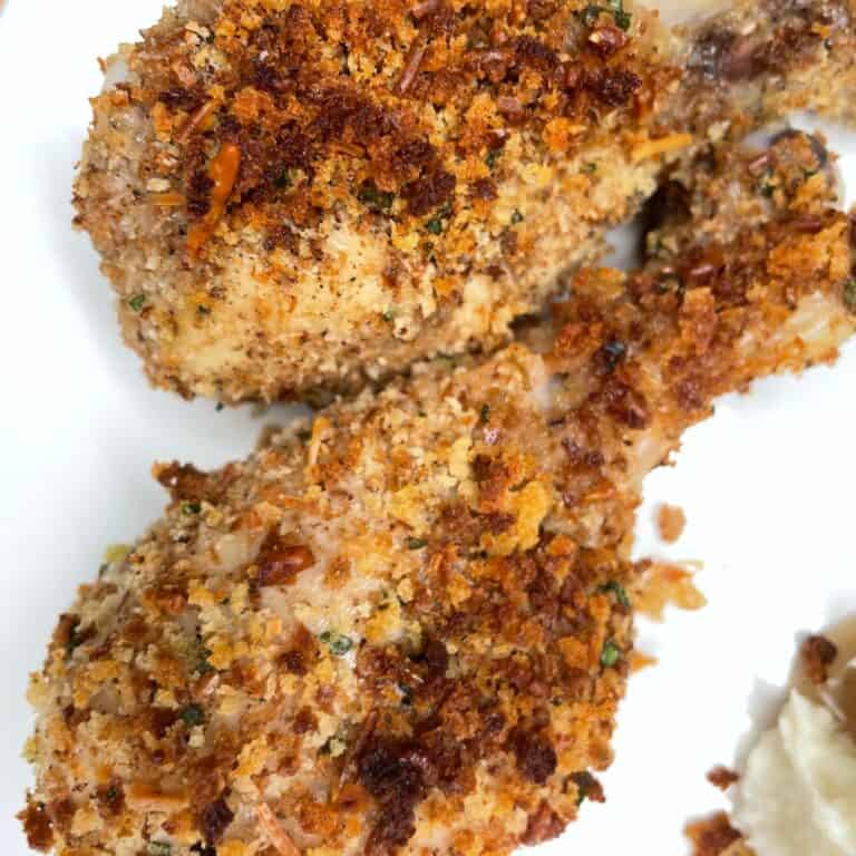 Two crispy, baked chicken drumsticks coated with a golden brown, crunchy breadcrumb and herb crust, served on a white plate. A small portion of mashed potatoes is visible at the edge.