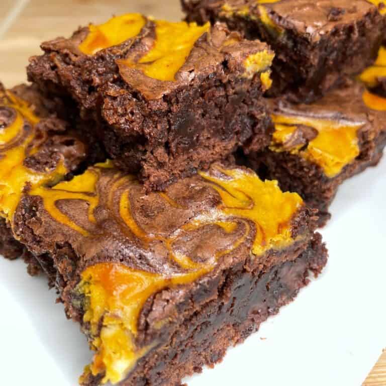 Close-up of rich, fudgy brownies with vibrant orange and yellow swirls on top, stacked on a white plate. The brownies have a moist, chocolatey texture and a marbled, colorful topping.